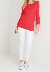 Rot gepunktete Langarmbluse mit einem Schlüsselloch-Detail am Ausschnitt, kombiniert mit weißen Slim-Fit-Hosen und weißen Sneakers.