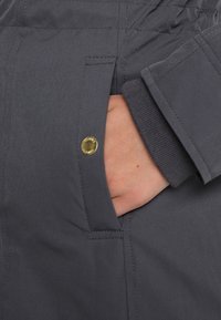 Main insérée dans la poche d'un manteau gris foncé avec un bouton-pression doré et des coutures visibles sur le tissu.