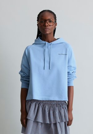 Frau mit geflochtenem Haar, die große braune Brille trägt, ein hellblaues Kapuzen-Sweatshirt und einen karierten mehrlagigen Rock, die vor einem schlichten hellen Hintergrund steht.