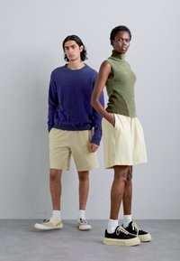 Dvaja modeli stoja vedľa seba. Mužský model má na sebe voľný, oversized fialový sveter a krémové šortky. Ženský model má na sebe priliehavý olivovo-zelený top bez rukávov a krémové šortky. Oboch zdobia biele ponožky so športovými topánkami.