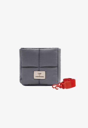 Graue, gesteppte, rechteckige Handtasche mit einem weißen Etikett vorne und einem abnehmbaren, breiten roten Träger mit Haken daneben.