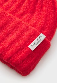 Red wool beanie with ribbed texture and a folded cuff, featuring a white tag with "Marc O'Polo" branding. Soft, warm material.
