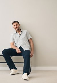 Camicia a maniche corte con motivo in colori chiari, pantaloni blu navy e sneakers bianche. Seduto su una sedia contro una parete neutra.