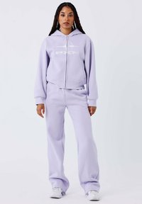 Lila rits hoodie met witte logo, gecombineerd met bijpassende sweatpants. Beide hebben een zachte, gestructureerde stof en een comfortabele pasvorm. Witte sneakers.