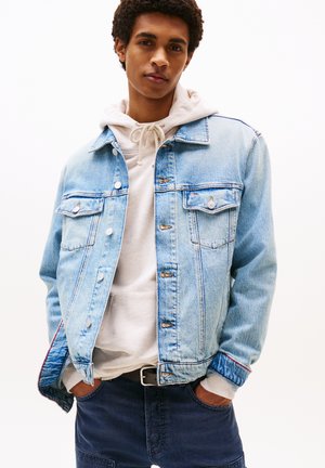 Chaqueta de mezclilla azul claro con dos bolsillos en el pecho, botones plateados y detalle de costura roja en los puños, llevada sobre una sudadera con capucha gris clara.