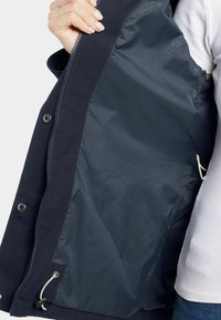 Marinblå yttertygsjacka med ett mönstrat mörkt foder, metallknäppningar och en justerbar dragsko i nederkanten.