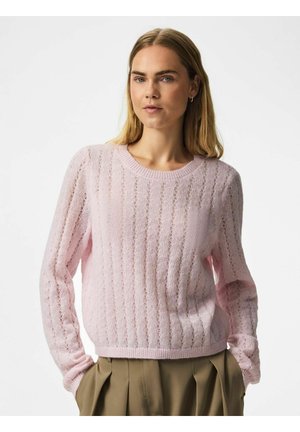 Femme aux cheveux blonds lisses porte un pull en maille rose clair et un pantalon plissé beige, debout les mains dans les poches devant un fond uni.