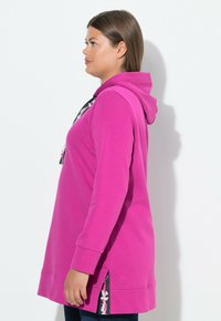 Felpa fucsia con cappuccio, maniche lunghe, spacchi laterali e dettagli grafici sulla parte anteriore e laterale. Texture morbida e liscia.
