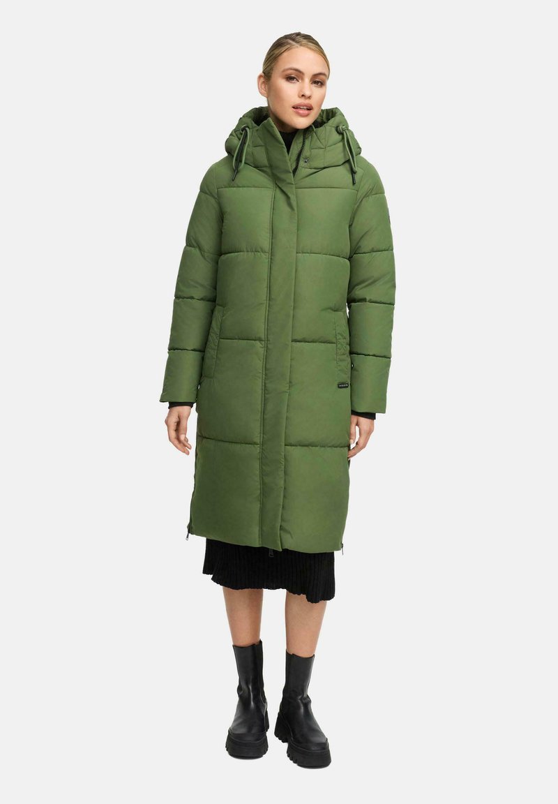 Kvinna i en lång grön vadderad kappa med huva, svart klänning och svarta ankelskurna stövlar, stående framför en neutral ljus bakgrund.