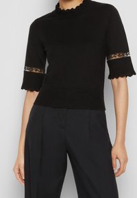 Top negro de punto con cuello festoneado y mangas cortas que presentan detalles de encaje transparente. Textura suave, diseño ajustado al cuerpo y acentos acanalados.