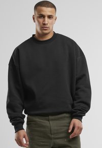 Felpa nera con scollatura rotonda, taglio ampio e maniche lunghe. Il tessuto appare morbido e texture, abbinato a pantaloni verde oliva.