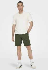 Camiseta polo de color beige claro con mangas cortas, combinada con pantalones cortos verde oliva doblados en el dobladillo. Zapatillas blancas con detalles en negro completan el look.