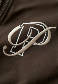Bruine stof met een geborduurd 'P' logo in beige en zwart. Het ontwerp heeft een gebogen staart die van de letter uitsteekt. Zachte textuur.