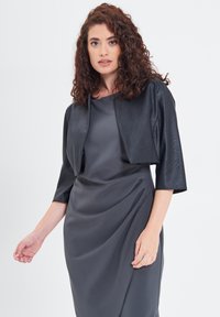 Giacca nera cropped in tessuto strutturato sopra un vestito grigio. La giacca presenta maniche ampie e un design pulito e minimalista.