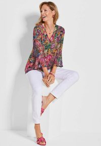 Bluse mit Blumenmuster und Glockenärmeln, überwiegend in Pink und Grün. Getragen mit weißen Hosen und rosa Riemchensandalen, sitzend auf einem weißen Podest.