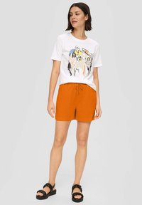 Witte t-shirt met een kleurrijke graphic print en oranje shorts met een elastische tailleband. Zwarte, chunky sandalen met gevlochten banden.