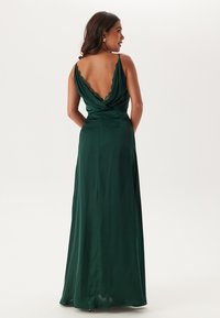 Abito in satin verde lungo fino ai piedi con scollatura profonda sulla schiena, dettagli drappeggiati sulle spalle e una silhouette fluida e morbida.