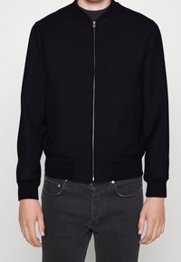 Giubbotto bomber nero con zip frontale, polsini e orlo a costine, realizzato in un tessuto liscio. Include tasche laterali e collo rotondo.
