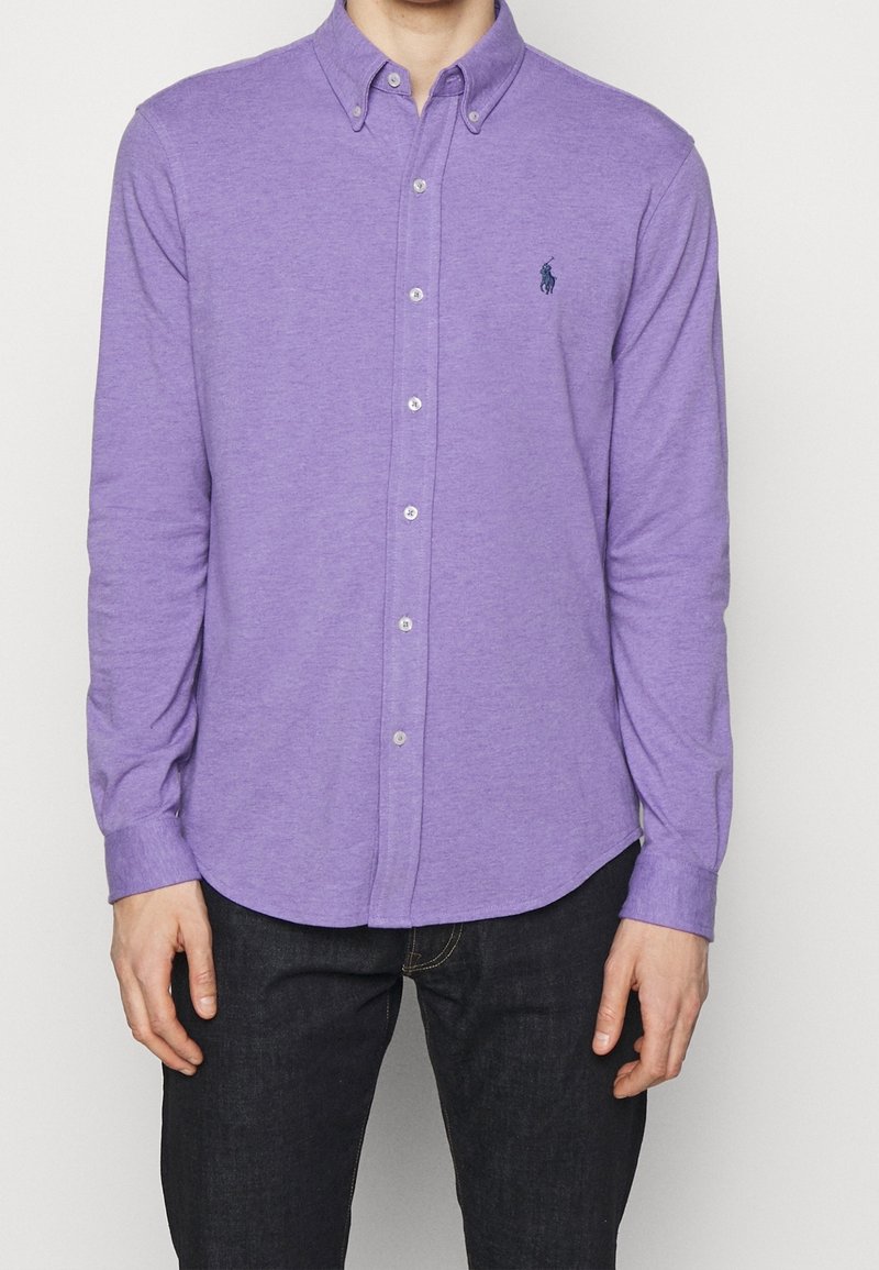 Fialová košile s dlouhým rukávem a límečkem, s jemnou texturou a malým vyšitým logem na hrudi.
