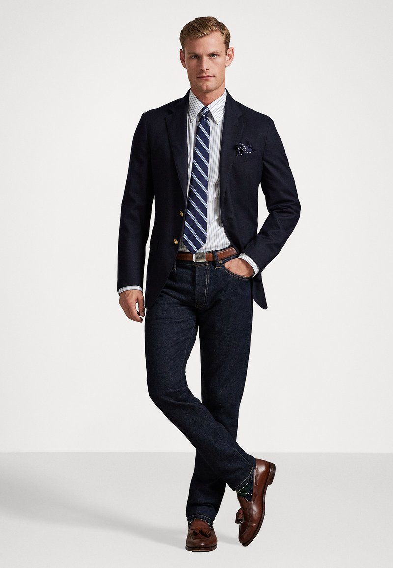 Navy blazer over a striped shirt with blue and white tones; dark jeans and brown leather loafers with decorative accents. Pocket square present.