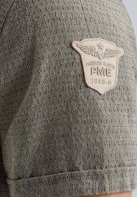 Siva majica s kratkimi rokavi z strukturirano tkanino. Vključuje kremno obarvan vezen logotip z krili in besedilom "PME 1549-A."