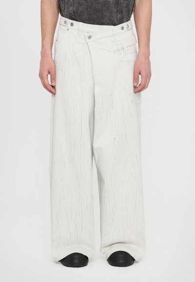 Pantalones de mezclilla blancos de corte oversized con una textura de líneas en espiral, cierre de botón y broche, y bolsillos laterales. El calzado tiene un acabado negro mate.
