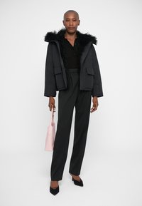 Giacca nera imbottita con rifinitura in pelliccia, pantaloni neri lunghi e tacchi neri. Tiene in mano una borsa rosa chiaro. Design semplice e moderno.