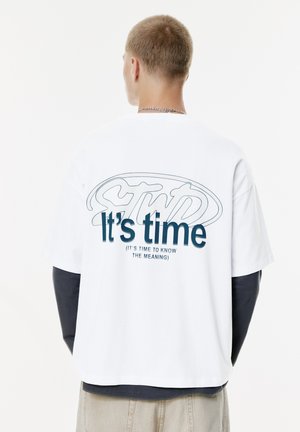 Ung mand med kort hår iført en hvid T-shirt over en langærmet sort skjorte, tekst på ryggen lyder "Det er tid (Det er tid til at kende betydningen)"