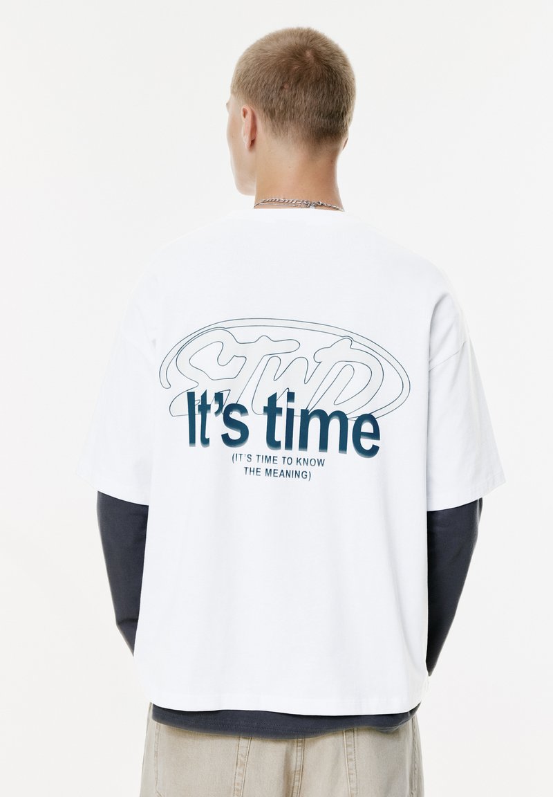 Ung mand med kort hår iført en hvid T-shirt over en langærmet sort skjorte, tekst på ryggen lyder "Det er tid (Det er tid til at kende betydningen)"