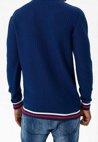 Maglione navy lavorato a maglia con un motivo testurizzato, caratterizzato da polsini e orlo a coste con strisce rosse e bianche. Indossato sopra dei jeans blu.
