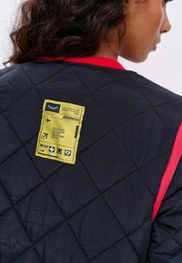 Osoba v černé prošívané bundě s červeným lemem a žlutou nášivkou s nápisem "Destinace: Svět, kde karikatury a dobrodružství nikdy nekončí."