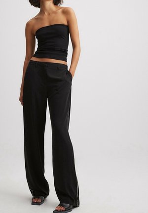 Mujer con un top negro sin tirantes y pantalones negros sueltos con detalles laterales, de pie con una mano en el bolsillo frente a un fondo liso.