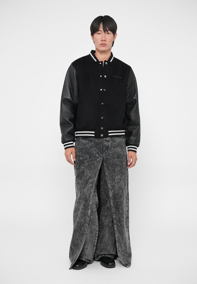 Giacca varsity nera con maniche in pelle, strisce bianche sui polsini, abbinata a pantaloni in denim grigio a spacco con una texture sbiadita.