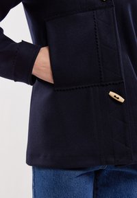 Marinblå ullblandad jacka med en stor ficka, vågig nederkant och träknapp. Synlig sömnad ger struktur och detalj till designen.