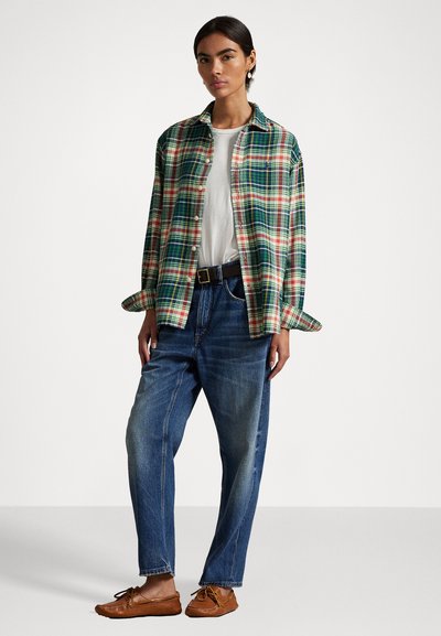 Kvinne som har på seg en grønn og rød rutete skjorte over en hvit t-skjorte, løse blå jeans, svart belte og brune mokasiner, stående på en ensfarget bakgrunn.