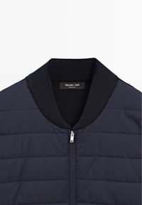 Navy quilted jacket with a ribbed collar and zipper closure. Features textured panels and a label inside the collar.