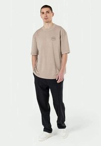 Béžové oversized tričko s okrúhlym výstrihom a malým logom, v kombinácii s čiernymi voľnými nohavicami a bielymi teniskami.