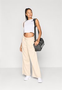 Fehér croptop fekete logóval, bézs bő szárú melegítőnadrág, és fehér sportcipők. Fekete táskát tart, amelyen fehér márkajelzés látható.