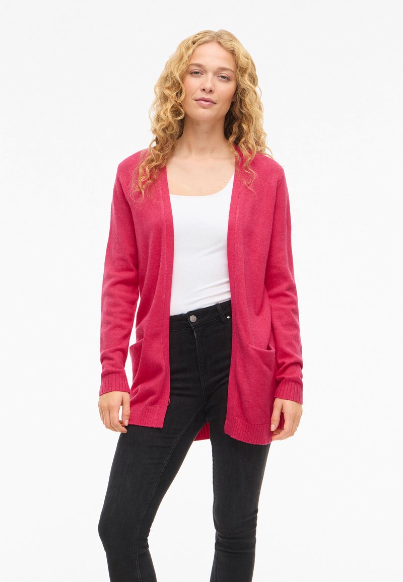 Long pink cardigan with open front, ribbed cuffs, and two front pockets. Worn over a white top and paired with black jeans.