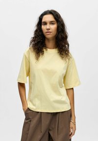 Femme aux cheveux bouclés et foncés, debout les mains dans les poches, portant une chemise jaune pâle à manches courtes et un pantalon marron ample, sur fond blanc.