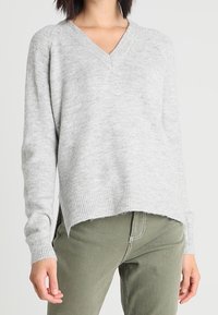 Šedý svetr s v-neck výstřihem vyrobený z měkkého pleteného materiálu, s uvolněným střihem, žebrovaným lemem a mírně delším zadním dílem. V kombinaci se zelenými kalhotami.