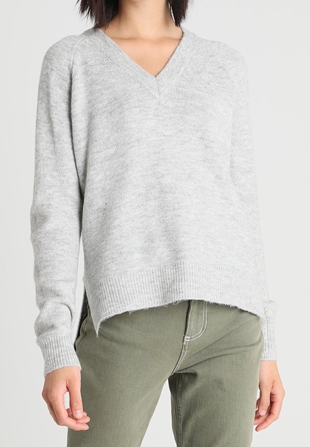 Šedý svetr s v-neck výstřihem vyrobený z měkkého pleteného materiálu, s uvolněným střihem, žebrovaným lemem a mírně delším zadním dílem. V kombinaci se zelenými kalhotami.