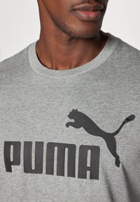 Grå bomull t-skjorte med en stor svart "PUMA" logo og en hoppende pantergrafikk over. Rund halsdesign med korte ermer.