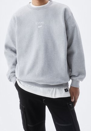 Person iført lys grå sweatshirt med lille hvidt logo tekst, lag-på-lag over hvid skjorte, kombineret med sorte cargobukser med synlige sømme.