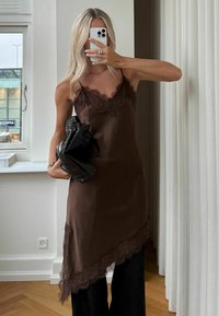 Pikkade blondide juustega naine, kes kannab pruuni pitsiga ääristatud rihmkleiti ja musti pükse, hoides kootud musta käekotti ja poseerides peeglis selfipilti tegemas.