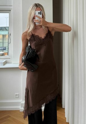 Pikkade blondide juustega naine, kes kannab pruuni pitsiga ääristatud rihmkleiti ja musti pükse, hoides kootud musta käekotti ja poseerides peeglis selfipilti tegemas.