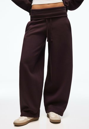 Person iført mørkebrune, vidde sweatpants med snøre i taljen og beige sneakers, stående mod en ensartet baggrund.