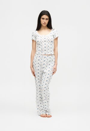 Jonge vrouw staat blootsvoets, draagt een witte korte mouwen top met bloemenprint en bijpassende lange broek, tegen een effen lichte achtergrond.