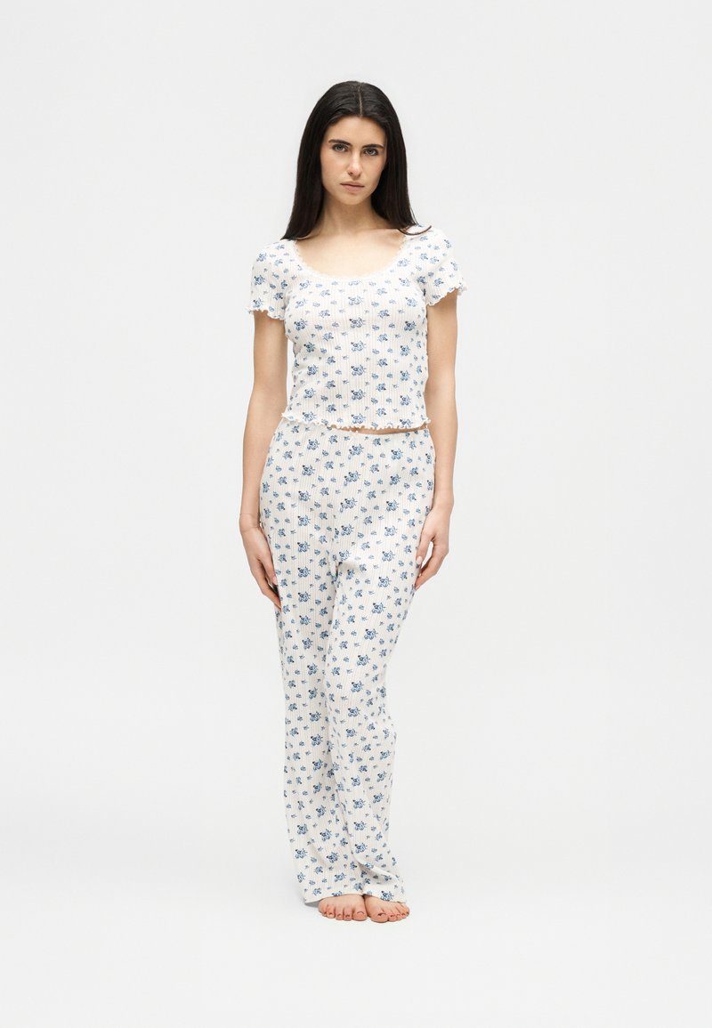 Νεαρή γυναίκα, ξυπόλυτη, φοράει λευκό φλοράλ τοπ με κοντά μανίκια και matching μακριά παντελόνια, μπροστά σε απλό ανοιχτόχρωμο φόντο.