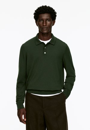 Dunkelgrünes Poloshirt mit langen Ärmeln und strukturierter Oberfläche, ausgestattet mit einer Knopfleiste mit drei Knöpfen und gerippten Bündchen, kombiniert mit dunklen Hosen.
